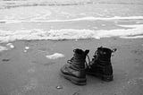 A black-and-white photograph of an empty pair of black shoe-boots at the edge of calm ocean water at the shore