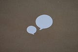 Two voice bubbles cut out of white paper on a brown background. One is small and the other large to illustrate the idea of the copywriting technique of starting short.