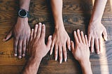 Picture of a wooden table with four peoples’ hands on side by side.