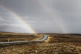 Photo of a road winding through open moorland with a grey sky and two rainbows in the distance.