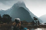 girl wearing toque sitting holding her legs looking at mountain across lake