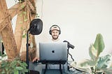 A podcaster or YouTuber sitting on his desk with his headset, his MacBook and green plants around him