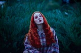 A white woman with David Bowie-esque bright pink and red makeup and long orange curly hair, wearing a black, pink, and white flannel and standing before a fern/bush.