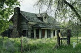 An abandoned house with dark windows and overgrown grass rising up the walls.