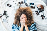 A woman lying on her back on a bed. She is surrounded by family photographs and two cameras. Some of the photographs show two people kissing. She has her hands over her face and appears to be in some distress.