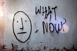 Graffiti with staid face and “What Now?” spray painted on the the wall of an abandoned building.