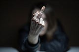 A person sits on a table and chair and shows the ‘middle finger’ to the camera, which also masks their whole face.