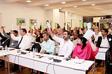 A group of mostly white people raise white slips of paper. In the front row, two brown people are not raising papers, and appear unhappy.