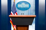 Image of a podium next to a US flag with the White House emblem behind it.