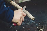An image of someone holding a small knife with a sharp tip whittling off the bark of a wooden stick.