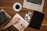 person writing in a journal in front of a laptop