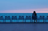 A woman sitting alone among empty chairs facing the ocean at sunset, looking out quietly