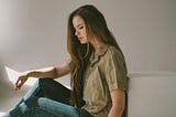 A woman with long brown hair and a contemplative look on her face, sitting in a stark white room by herself.