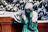 A green statue of a woman covered in snow and resting her head on one hand in grief or sadness, her other arm draped over a collumn attached to a brown wall, with leafy, snow-covered branches of a tree filling the background behind the brown wall.