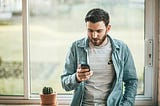Gentleman in gray tshirt with denim overshirt leaning on a window sill checking Instagram on his phone.