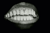 A mouth with lips, teeth and tongue speaking dramatically against a black background