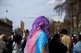Person with medium-long wavy light purple hair and the trans flag draped over their shoulders in a crowd