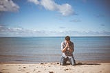 A man kneeling in prayer in front of a body of water.