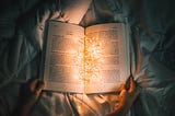 An open book on a blanket in a bed with lights inside the book, illuminating the pages