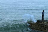 A man standing at the end of a pier with waves crashing against the rock wall