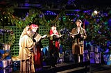 Three Carolers with song books in hand and decrative lights in the background.