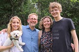 A young woman holding a small white dog, a middle aged man with glasses and a beard, a middle aged woman, and a tall young man with glasses all smile at the camera