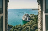 An arched window in a stone wall, looking out at the sea and boats