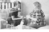 Two people sit in a corner, having serious conversation. One of them holds a clipboard