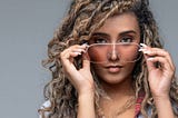 Studio portrait of an attractive and hip young woman with curly hair and sunglasses looking into the camera confidently
