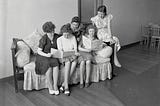 A black and white photo of women in dresses and skirts gathered on a couch reading pamphlets