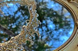 A sparkling royal crown sits atop a mirror that reflects trees and sky
