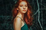 red haired woman in a green spaghetti strapped dress, staring at the camera. Behind here are pale branches against a dark background