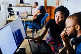 A young Black woman in a black shirt and colorful skirt pointing at a computer screen while talking to a young Black man with glasses and headphones, wearing a blue shirt. They are in an office with other employees visible in the background.