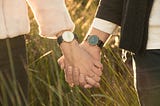 Two people holding hands in a field of grass, both wearing watches on their wrists.
