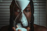 An hourglass full at the top with sand draining down is held by a woman who looks at it or us close up — closed horizontal window blinds are in the background.