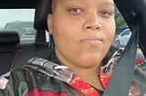 selfie of a light skinned Black woman in a car. She is looking at the camera with a blank expression. Her head is bald, and she’s wearing a hoodie that appears to have a red, brown, and gray pattern in the style of women’s armor.
