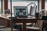 An open laptop computer sits on a kitchen table by a window overlooking a lake. A coffee cup and a book are beside it.