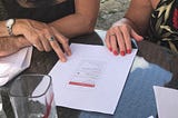 Photograph of a user research session, there is a paper prototype on the table with the hands of two women who are discussing their thoughts around the prototype.