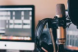 Headphones resting on a podcasting microphone with a computer screen blurred in the background.