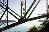 Steel bridge supports intersecting above calm water, framing open space beyond. A load-bearing structure holding tension while enabling movement beneath.