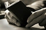 A very small, antique, hardback book is held open on a desk by the reader’s hands.