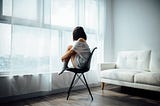 woman sitting on plastic chair in living room, looking out a large window, hugging her knees