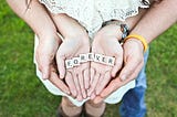 A picture of a parent holding a childs hands while the child holds the scrabble letters spelling “forever’