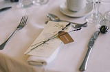 A dinner menu and fine silverware, atop a white-clothed table