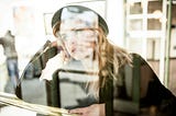 A young emerging adult wearing a black leather jacket and black hat leaning on her right arm looking out the window thinking — consulting with her future self.