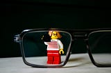 A lego woman wearing glasses peeks through a pair of glasses.