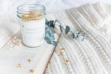 white candle called “manifest and chill” with eucalyptus and flower bundle sitting on top of an open book spread across a white blanket with gold stars splayed on top