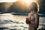 A white topless man in front of a sunset at a beach, his hands forming a mantra.