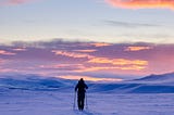 A man with skis walking through snow with the sunset in the background