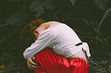 A person wearing a white long-sleeve top and red pants crouches on the ground in a forest, head down and arms wrapped around their knees, surrounded by fallen leaves and dark green foliage, conveying a mood of sadness or introspection.